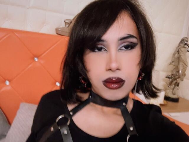 FemCupid with dark lipstick, black eye makeup, black choker, and short black hair, wearing a black top, sitting in front of an orange headboard.