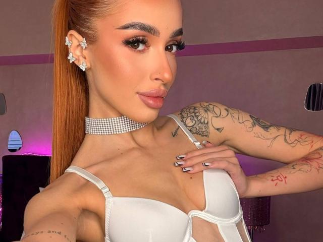 MarissaFex with long orange ponytail, wearing white bra, diamond choker, star earrings, tattoos on arms, hand on chest, pink lipstick, glossy nails, and sexy, confident expression.