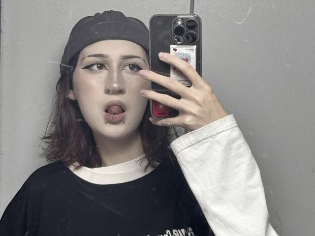 JeaneneSmolik with freckles, black baseball cap, white long-sleeve shirt, black tee, tongue out, septum piercing, and holding phone in mirror selfie, showing off her sexy face.