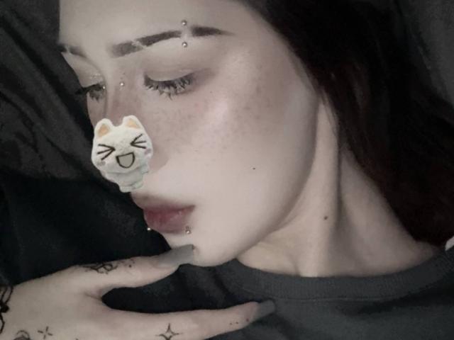 JeaneneSmolik has pale skin, dark brown hair, and multiple facial piercings. She's wearing a cute cat sticker on her nose, with her eyes closed and a hand near her mouth.