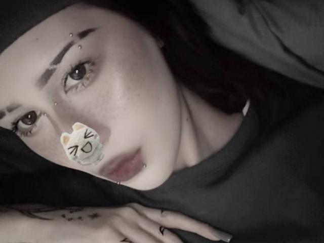 JeaneneSmolik's face, pale skin with dark eyebrows, eye piercings, nose boop sticker with cat ears, lip piercing, black shirt, tattooed hand resting on bed, dark hair.