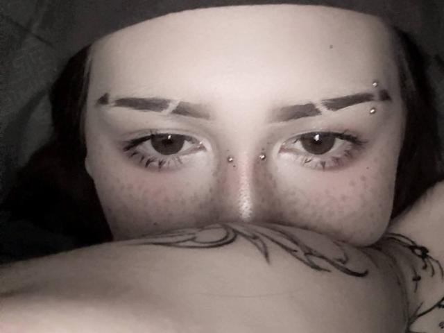 JeaneneSmolik's face with dark eyebrows, eye piercings, freckles, and tattooed shoulder, looking up with half-visible tits in the bottom frame. Her eyes are the focal point.