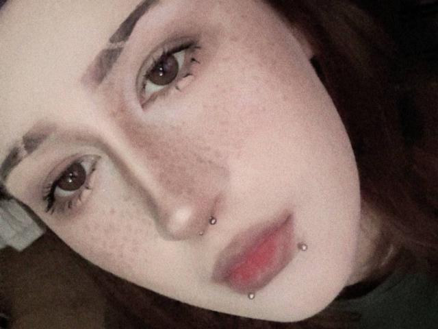 JeaneneSmolik's face with freckles, brown eyes, dark eyebrows, nose piercing, and lower lip piercing. She has red lips and brown hair. Her skin is pale.