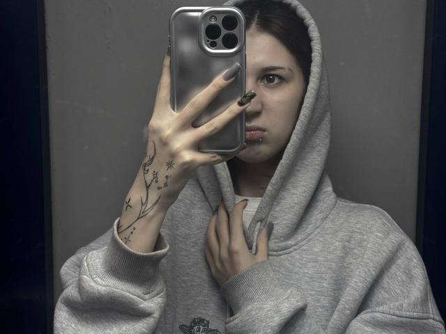 JeaneneSmolik with gray hoodie, holding silver iPhone, showing tattooed hand with black nails, looking at mirror, serious expression, gray background.