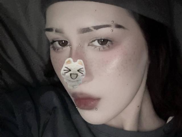 JeaneneSmolik's face has a cute cat sticker on her nose, pierced eyebrows, and a pierced lower lip. She's wearing a dark headscarf and a grey shirt.