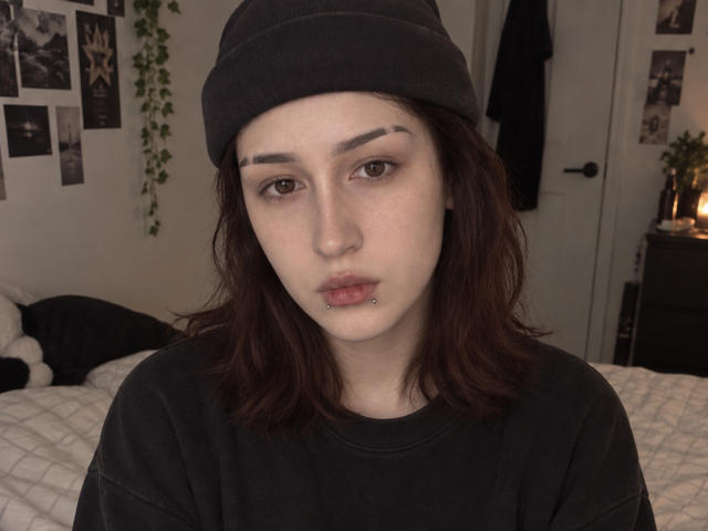 JeaneneSmolik has fair skin, dark brown wavy hair, black beanie, black shirt, and a lip piercing. She's looking straight with a neutral expression. Her tits are small.