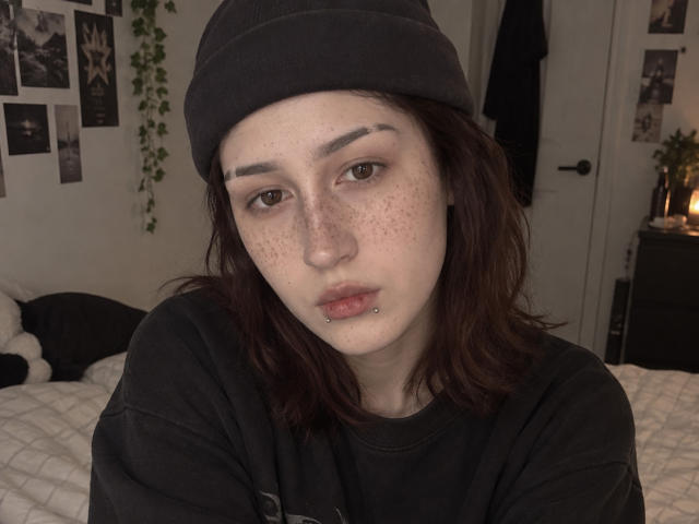 JeaneneSmolik with freckles, dark brown wavy hair, black beanie, black shirt, lip piercing, and brown eyes, looking at camera with slightly pouty lips.