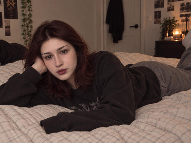 JeaneneSmolik, with pale skin and dark red hair, lies on a bed, wearing a black sweatshirt and gray pants, head resting on her hand, looking at the camera.