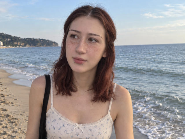 JeaneneSmolik with reddish-brown hair, freckles, and a nose piercing, wearing a white floral camisole, standing on a beach with small waves, black strap over shoulder.
