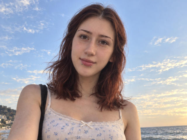 JeaneneSmolik with red hair, wearing a white floral tank top, nose ring, and lip piercing, standing by the ocean at sunset, holding a black strap.
