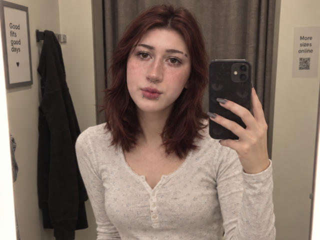 JeaneneSmolik with red hair, nose ring, and lip piercing, wearing a white long-sleeve shirt, taking a mirror selfie with a black phone, showing her tits.