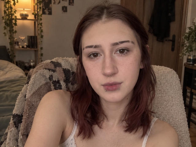 JeaneneSmolik has red hair, brown eyes, and a nose piercing. She's wearing a white tank top, and has a lip piercing with her arms crossed. Bedroom setting.
