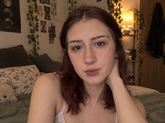 JeaneneSmolik with red hair, piercings in nose and lip, freckles, looking at camera, hand on neck, wearing white tank top, in dimly lit bedroom.