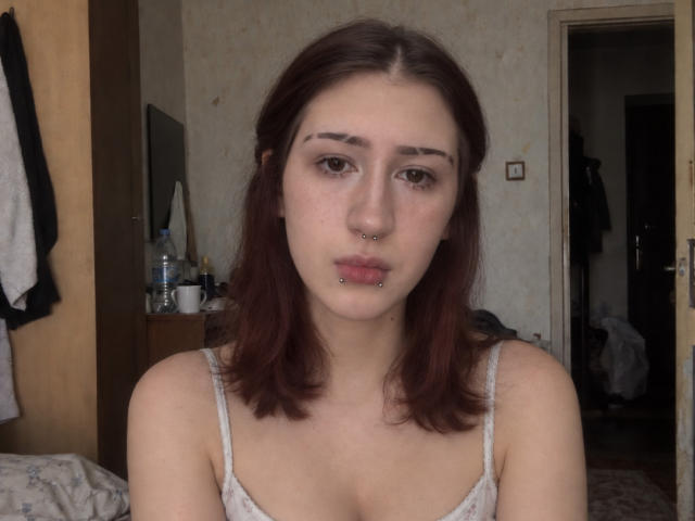 JeaneneSmolik has red hair, a nose piercing, a lip piercing, and is wearing a white tank top, showing her small tits and a neutral face expression in a bedroom.