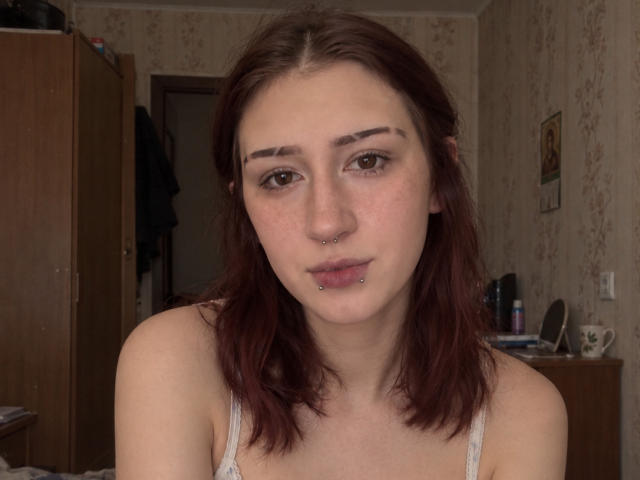 JeaneneSmolik has brown eyes, a nose ring, and lip piercings. She has freckles, dark red hair, and is wearing a white tank top, showing her bare shoulders.
