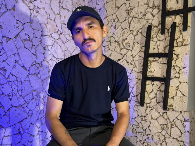 EmmaAndRick, short mustached dude with black cap, black t-shirt, and black pants, sitting with hands on knees, textured wall with ladder in background, slight blue lighting.