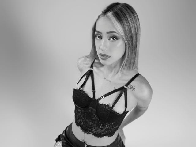 Kloepraga with blonde bob, black lace bra, strappy design, tattoo on right shoulder, looking up, hands behind back, gray background, black-and-white photo. Boobs, tattoo, strappy bra.