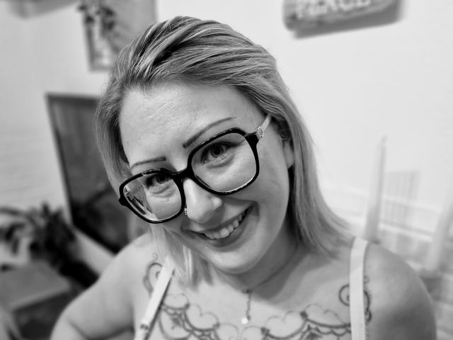 AmandalaFrancaise with glasses, smiling, showing tattoos on her chest, blonde hair, and a necklace, in a black-and-white photo. She's got a cute, nerdy, sexy look.