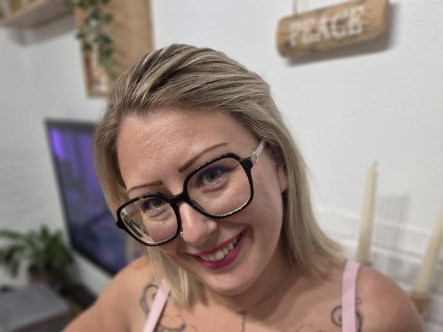 AmandalaFrancaise with blonde hair, black glasses, pink top, smiling, showing tattoos on shoulders, pink lipstick, nose piercing, white wall background, candles on right.