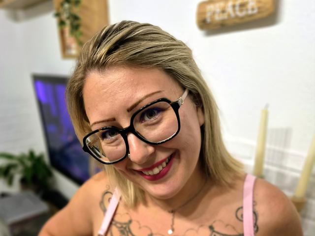 AmandalaFrancaise with blonde hair, black glasses, pink top, smiling, showing tattoos on chest, pink lipstick, nose piercing, white wall in background, blurred TV and plants.