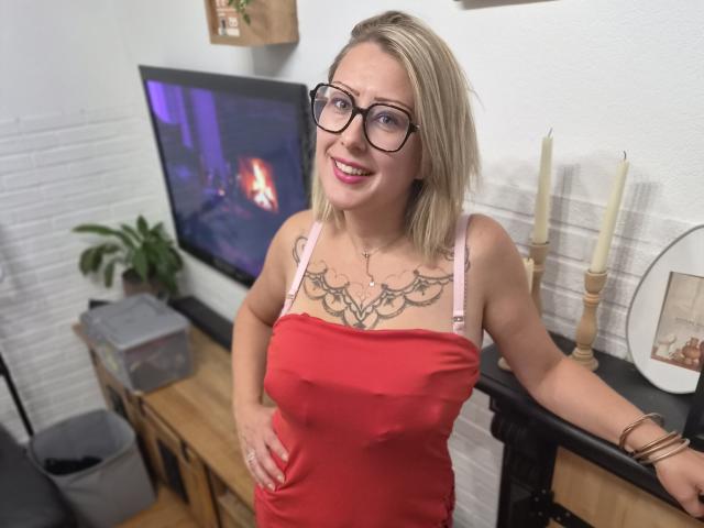AmandalaFrancaise with blonde hair, glasses, and pink straps, wearing a red tank top, showing tattoos on her chest, smiling, standing in a room with candles.