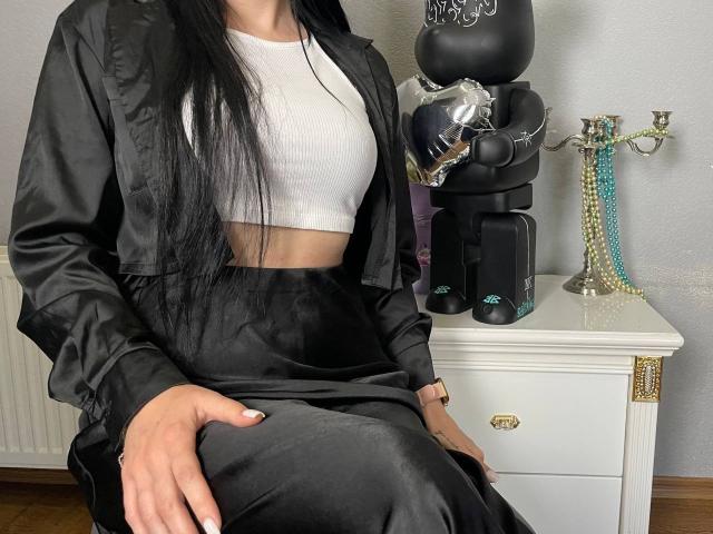 Xioni with long black hair, wearing a black satin jacket and high-waisted black satin pants, sitting on a white chair, white crop top showing her tits. Coffee maker and candelab on side.
