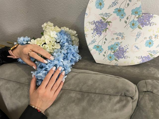 Xioni's hands resting on a gray couch, holding a bouquet of blue and white hydrangeas, with a floral-patterned pillow to the right. Xioni wears a gold watch and bracelet.
