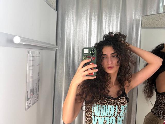 MyeshaDegennaro with curly brown hair, wearing a leopard print top, holding a green phone, taking a mirror selfie, hand in hair, looking at camera, in a bathroom.
