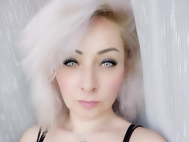 IlonaCharmy, com cabelo rosa claro, olhos azuis e delineador escuro, olhando diretamente para a câmera, vestindo uma blusa preta e um colar de prata, com uma aparência fofa e levemente emburrada..