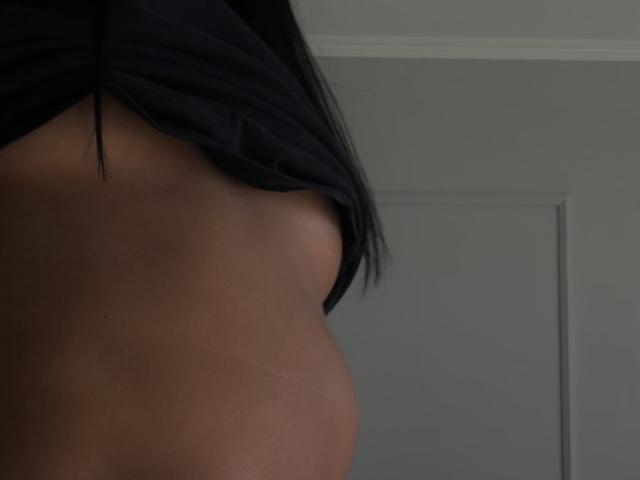 Fridaa's bare side with a raised black shirt, showing a smooth brown breast and part of her black hair, in a dimly lit room.