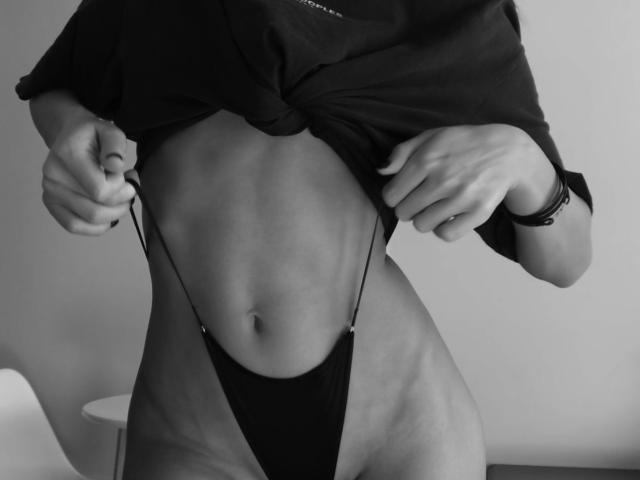 Black-and-white photo of Fridaa lifting a shirt, revealing a slim, toned belly and wearing a tiny black thong that accentuates her pussy. She's pulling the shirt up with both hands.