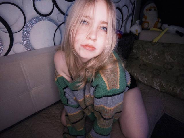 TiffaniMacandog, blonde with pale skin and blue eyes, wearing an off-shoulder green and gray striped sweater, kneeling on a bed with green pillows. Her pussy is not visible.