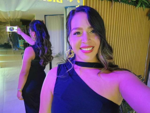 LiaDiane with wavy brown hair, smiling, wearing a black one-shoulder dress, holding a camera in the background, purple and yellow lighting, large gold earrings, visible butt.