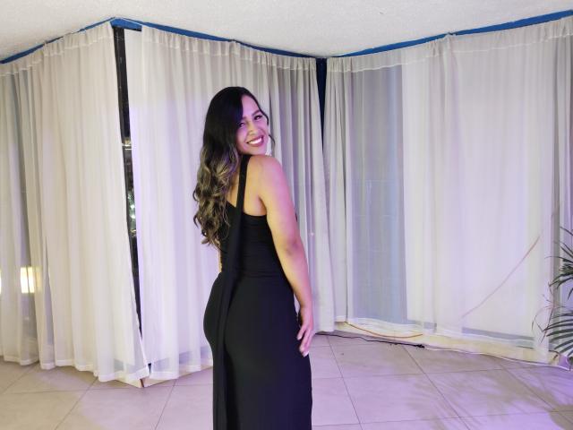 LiaDiane with long brown hair, smiling, wearing a black dress, showing her ass, standing in a room with white curtains and a tiled floor.