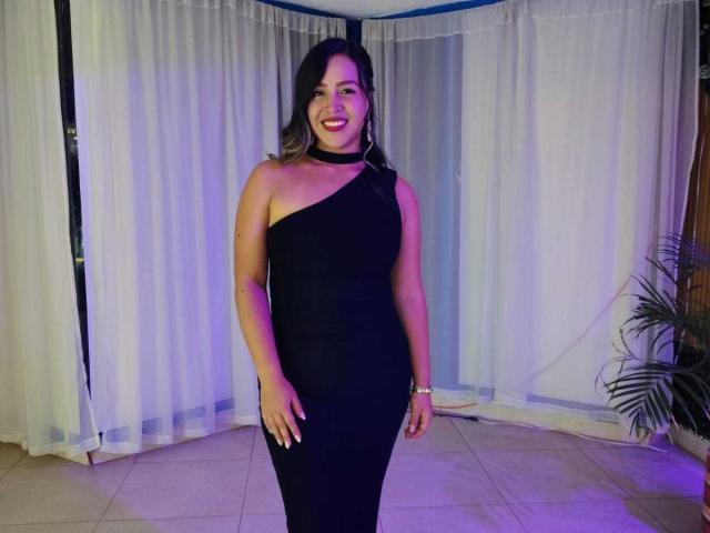 LiaDiane, a curvy woman with black hair, wearing a black one-shoulder dress, smiling with red lipstick, standing in front of white curtains, showing her ass.