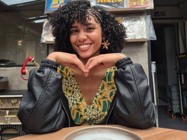 Cinthia Walker mit lockigem, schwarzem Haar, lächelnd, mit Händen, die eine Herzform bilden, trägt ein grünes und goldenes Oberteil und eine schwarze Lederjacke und sitzt an einem Holztisch..