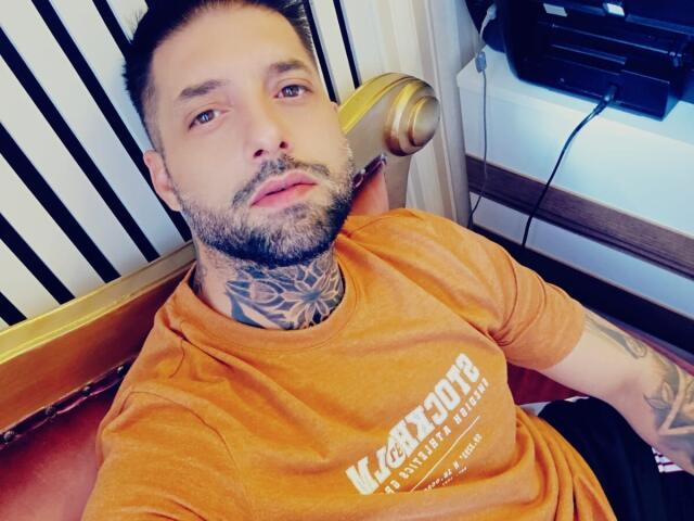 TommyDum has a beard, tattoos on his neck and arm, and is wearing an orange shirt. He's sitting with a relaxed, slightly annoyed expression.