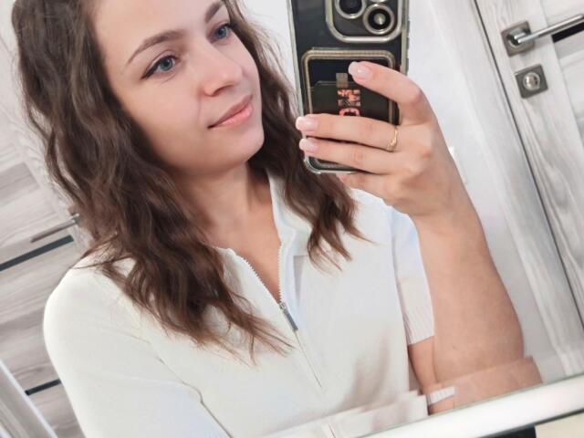 SaraUili with wavy brown hair, light skin, and small breasts, wearing a white zip-up shirt, taking a selfie in a mirror with a black phone.