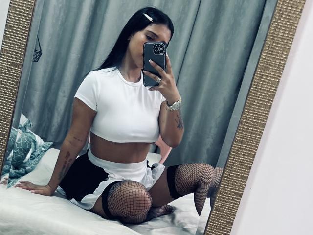 SophieCan, vestida con un top corto blanco, una falda negra y medias de rejilla, está sentada en una cama, tomando una foto de sí misma con su teléfono en un espejo, mostrando su trasero y sus tatuajes..