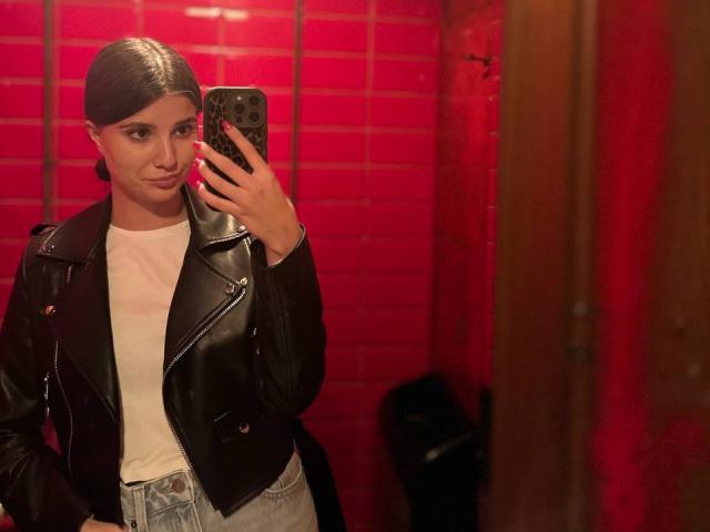 LeiaLindman with slicked-back brown hair, black leather jacket, white tee, and high-waisted jeans, taking a mirror selfie, holding a black phone, red-lit bathroom.
