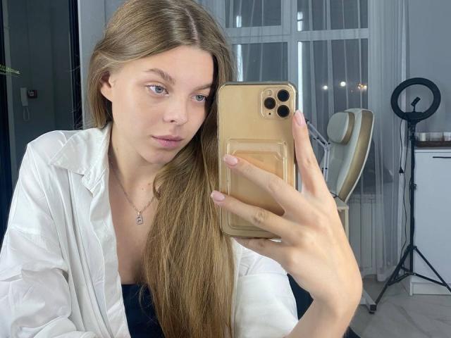 RhonaBells with long blonde hair, wearing a white shirt, taking a selfie with a gold iPhone, showing her small tits and pale skin, in a bright room.