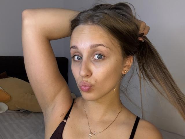 TrissyTerry with puffy lips, raising arm, brown hair in ponytail, black tank top, small gold earrings, necklace, light skin, green eyes, slight tattoo on upper arm.