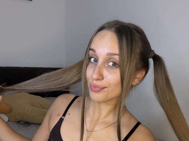 TrissyTerry with pigtails, blue eyes, pink lips, black spaghetti strap top, small tits, light skin, holding hair, gold necklace, slight smile, in a bedroom.