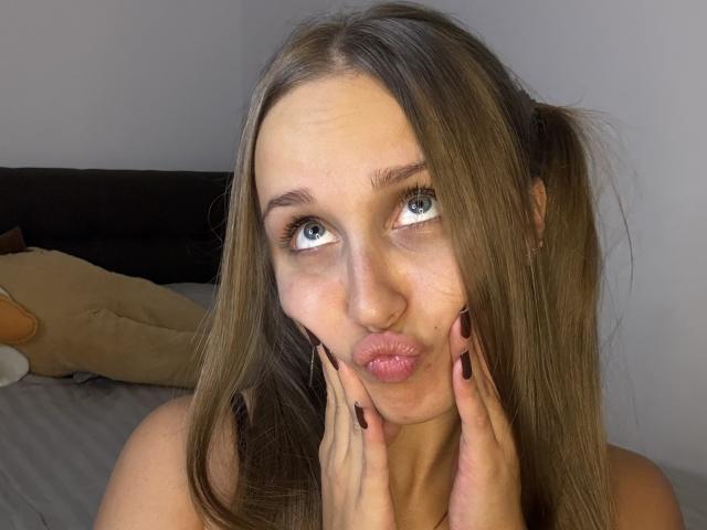 TrissyTerry pouts her pink lips, hands on cheeks, with long brown hair, green eyes, and black nail polish, in a selfie. Her tits are not visible.