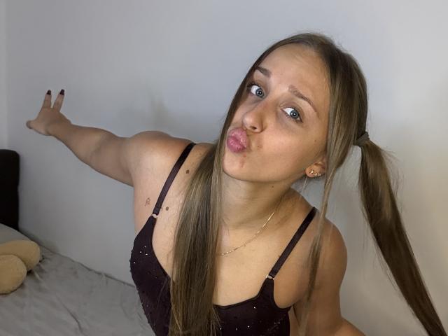 TrissyTerry with puffy lips, blue eyes, and long brown pigtails, wearing a black sparkly top, reaching out with a raised arm, showing her small tits and tattoos.