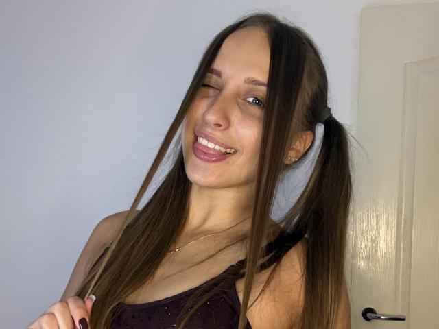 TrissyTerry with long brown pigtails, winking and smiling, showing her teeth, wearing a dark purple top, in a room with a white door. Her tits are small.