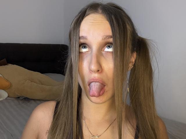 TrissyTerry has long brown pigtails, blue eyes, and her tongue out. She's wearing a necklace, and her face is slightly squished with a playful expression.