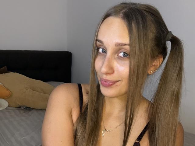 TrissyTerry with pigtails, wearing a black bra, has a cute smile and light makeup, showing her tits and small gold necklace, standing by a bed with a stuffed animal.