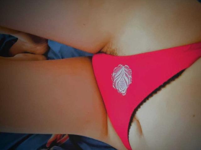 MissSunnyShine's pink thong with a white leaf design, sitting on her crotch, showing her tan-lined pussy and some dark pubic hair. Her hand is near her thigh.