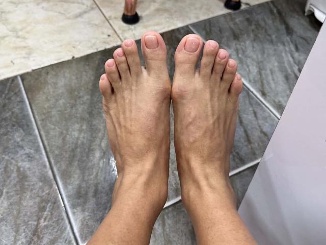 Los pies de JoseRumBerotop, mostrando dos dedos descalzos con lechos ungueales rosados, descansando sobre un suelo de baldosas grises, piernas ligeramente separadas, tobillos visibles, tono de piel marrón claro, definición muscular sutil..
